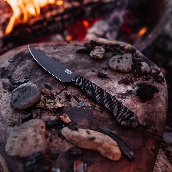 Montana Knife Company - Speedgoat Fixed Blade - Black - Parachute Cord - Knife laying on rock by fire surrounded by rocks and small pieces of wood and char.