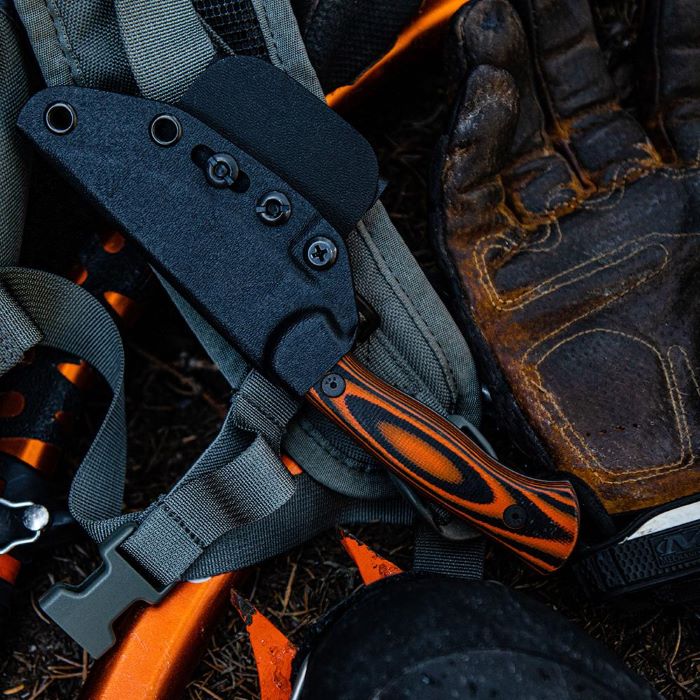 Montana Knife Company - The Blackfoot Fixed Blade 2.0 - Orange and Black - Knife in black kydex sheath attached to front strap of a backpack  that is resting on the groundn ext to a pair of outdoor gloves