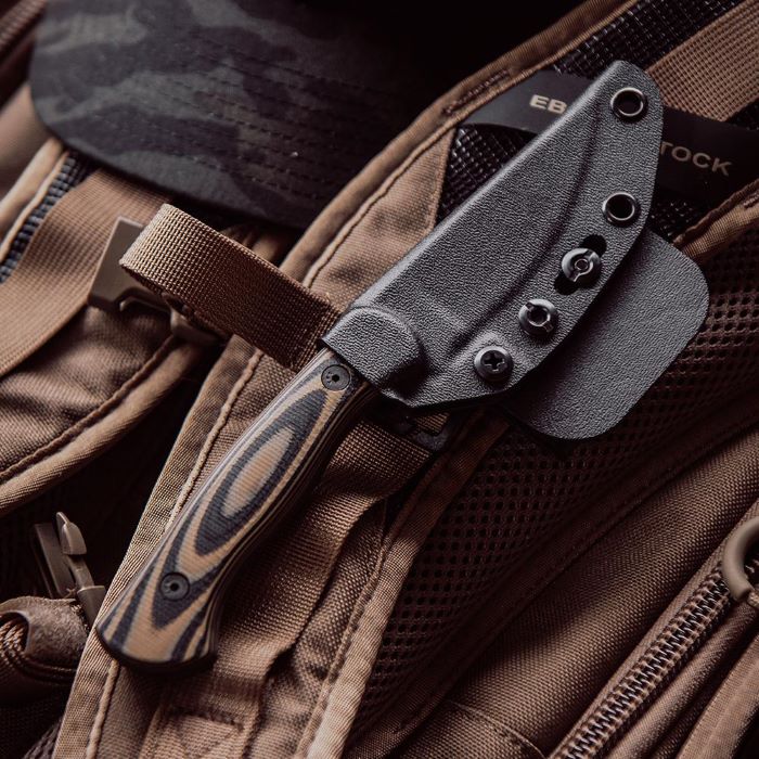Montana Knife Company - The Blackfoot Fixed Blade 2.0 - Tan and Black - knife in black kydex sheath attached to an eberlestock backpack strap resting on the ground next to a camo hat