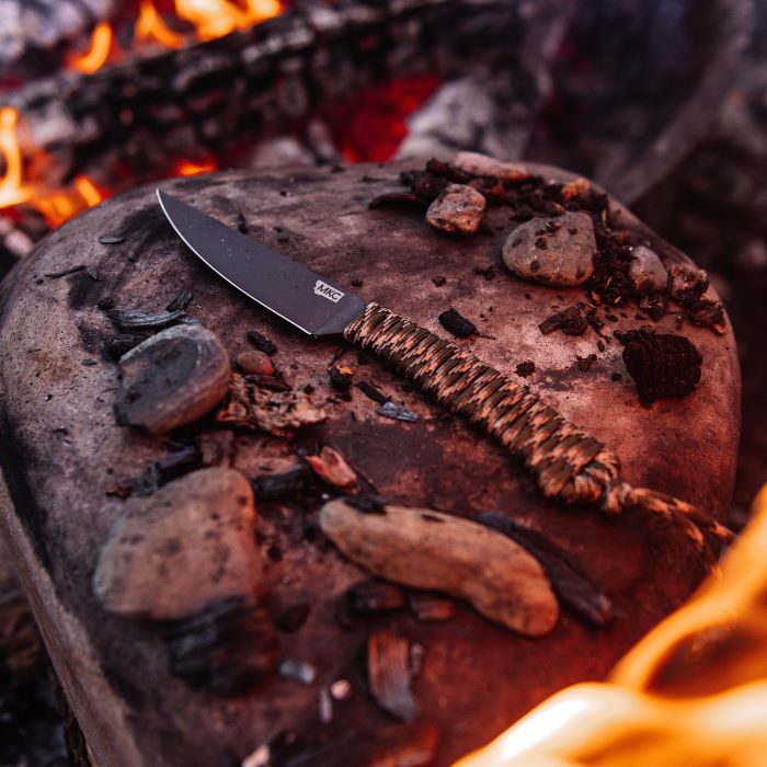 Montana Knife Company - The Speedgoat Fixed Blade - Forest Camo - knife laying on rock in front of fire next to small stones and charred wood pieces
