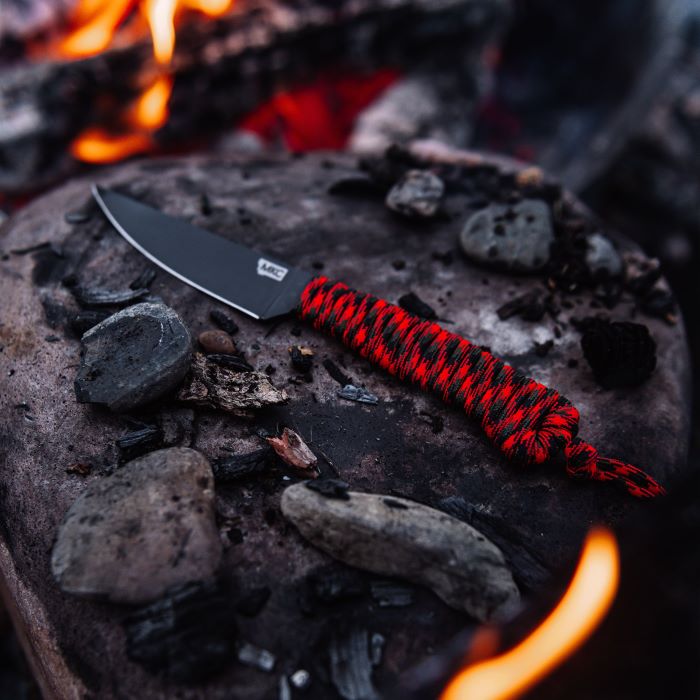 Montana Knife Company - The Speedgoat Fixed Blade - Blaze Orange and Black - Parachute Cord - Knife on rock in front of fire next to small stones and charred wood pieces