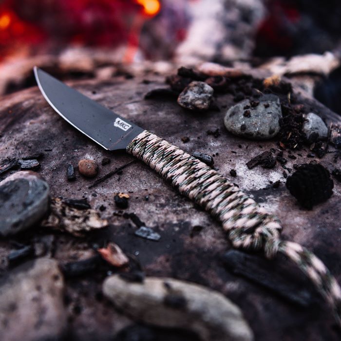 Montana Knife Company - The Speedgoat Fixed Blade - Desert Camo - Knife laying on rock in front of fire next to small stones and charred wood pieces